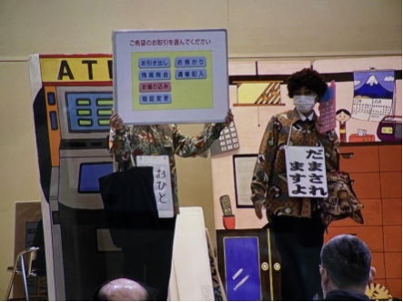前に2名の人が立ち、右の人が「だまされますよ」のカードを首に下げ、防犯教室を行っている写真
