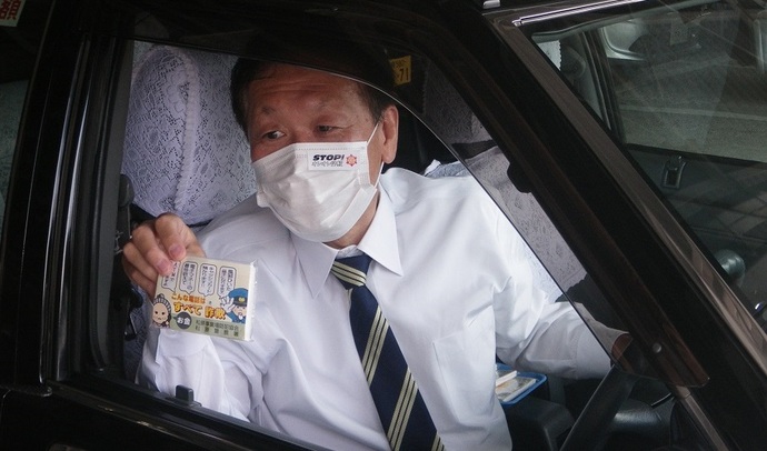 タクシー運転手さんが運転席から窓を開け、STOP!特殊詐欺のシールを貼ったマスクを着用し特殊詐欺啓発ポケットティッシュを左手で持っている写真