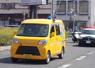「NPO法人やんちゃまファミリーwith」の黄色の車を先頭に、パトカー、警ら用の単車に乗った警察官が道路を走っている写真