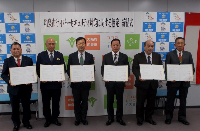 和泉市サイバーセキュリティ対策に関する協定締結式で6名の関係者の方々が協定書を持って1列に並び記念撮影をしている写真