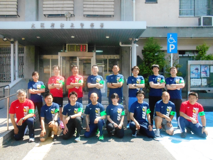 和泉警察署員の方々が赤と青の色違いでお揃いのTシャツをきて左腕には緑の腕章をはめ、2列に並んで記念撮影をしている写真