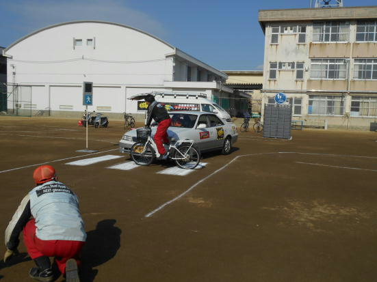 校庭に白線で道路と横断歩道を書き、信号機の模型を置き、横断歩道を自転車で渡っている所、右から走ってきた車と接触している写真
