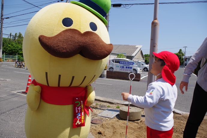 「大阪府貝塚市つげさん」と書かれた名札を左胸につけ緑の帽子に大きな口ひげが特徴の貝塚市マスコットキャラクター「つげさん」に話かけている体操着姿の園児の写真