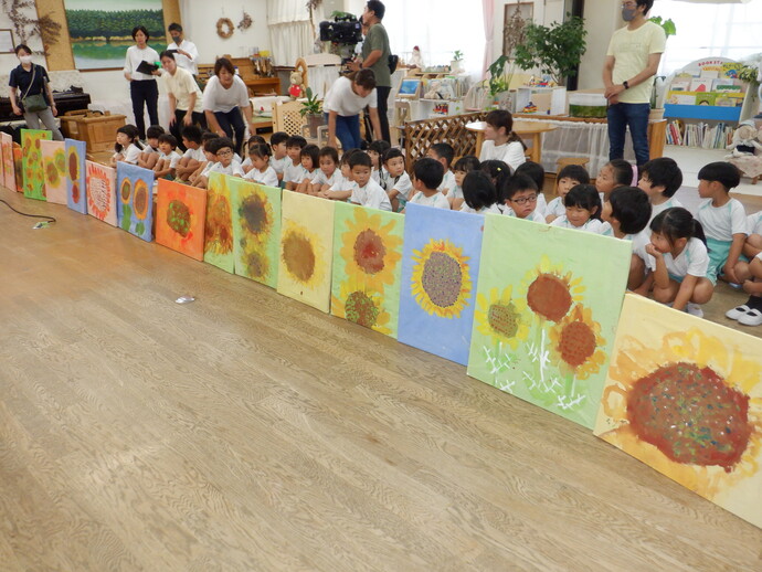 室内で、園児たちが2列ほどに並んで座り、前方に作成したひまわりの絵を並べて展示し、関係者に披露している写生大会の写真