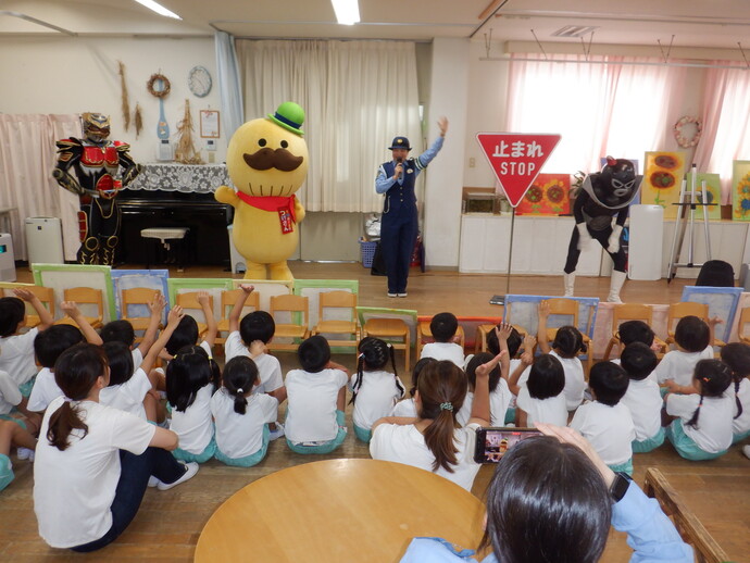 園児たちが作成したひまわりの絵が並べてある教室で貝塚市マスコットキャラクターの「つげさん」とご当地ヒーロー「浪速伝説トライオー」と悪役キャラクターがマイクを持ち片手を挙げた女性警察官が2列ほどに並んで座り同じく片手を挙げている園児たちに向けて話をしている交通安全教室の写真