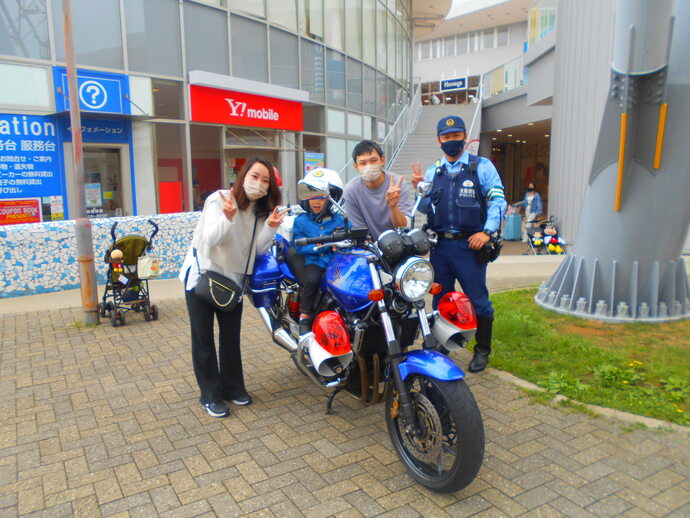 小さな男の子がヘルメットを被り青バイに乗り、左側に女性、右側に男性、警察官ら3人がピースしている写真
