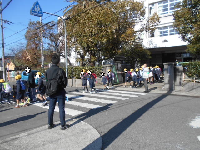 小学校の門の前の横断歩道を渡る生徒をパトロール隊が見守っている写真