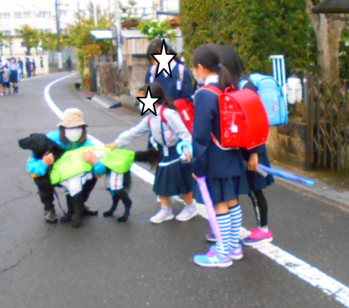 愛犬パトロール隊（わんだふるくらぶ）の黒い犬を、下校中の3人の女の子が見つめ、1人が背中を撫でている写真