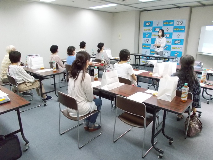 室内の前方に1人の女性が立ち、他の参加者は長机に設置された椅子に座りながら腕章授与式に参加している様子の写真