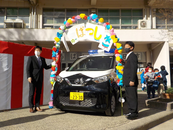 三日市小学校で、「しゅぱつしき」と書かれた紙の花を飾ったアーチの下に、青色防犯パトロールカーが停まっており両脇に関係者が立ち記念撮影している写真