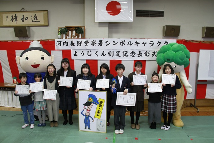 「河内長野警察署シンボルキャラクターようじくん制定記念表彰式」と書かれた横断幕の前で、表彰状を持った10名の子ども達を中心に、河内長野市シンボルキャラクターの「モックル」くんと河内長野市生涯学習推進マスコット「くろまろくん」が並んで記念撮影をしている様子の写真