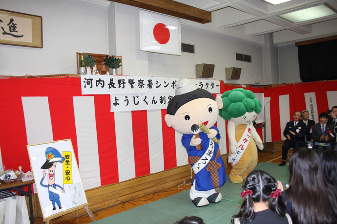 紅白の横断幕が張られた会場で、多くの関係者たちが着席しているなか、モックルくんと、くろまろくんが正面に立っている写真