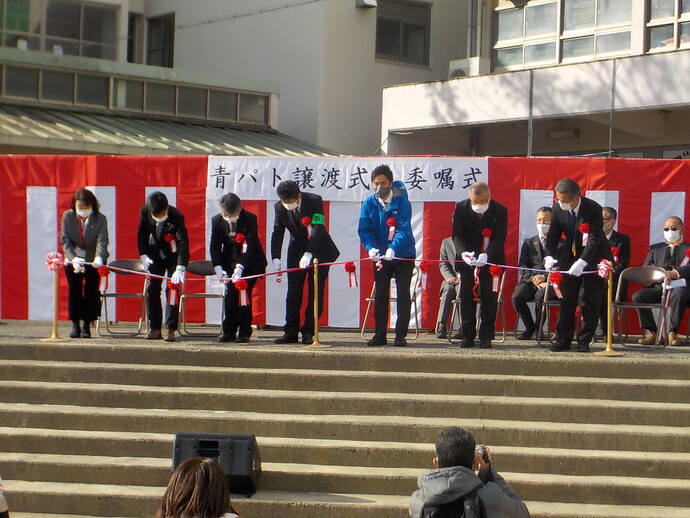 青パト譲渡式・委嘱式と書かれた紅白の横断幕の前で、7名の式典参加者がテープカットをしている写真