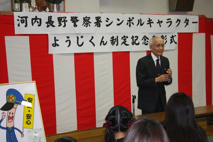 「河内長野警察署シンボルキャラクターようじくん制定記念表彰式」と書かれた横断幕の前で、稲葉会長がマイクを手に講和をしている様子の写真