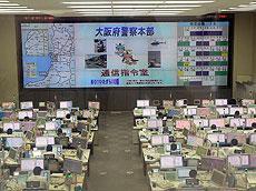 全面の大きな画面に大阪府の地図と大阪府警察本部通信指令室と映し出されている通信指令室全景（見学者コーナー）
