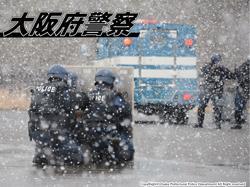 吹雪の中、機動隊の車輌と手前には盾を構える2人の機動隊員の写真