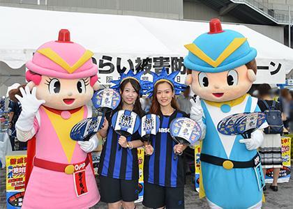 大阪府警察ブースにて、ガンバ大阪のユニフォームを着た女性と警察のキャラクターがうちわを持って並んだ写真