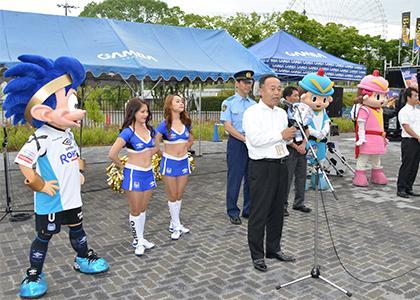 ガンバ大阪や警察のキャラクター、警察官やチアリーダーが並び、男性が挨拶をしているセレモニーの様子