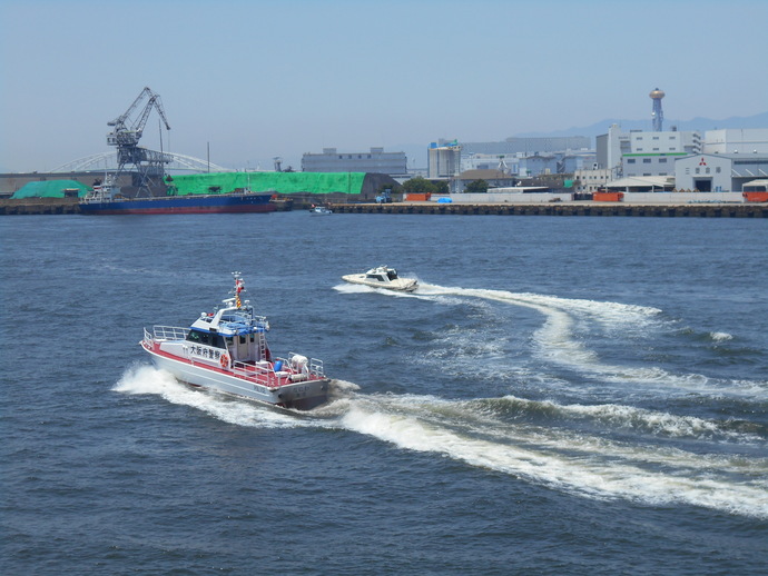 阪港港天保山岸壁の海上で大阪府警察と書かれた警備艇が波しぶきをあげている外国貨客船を追跡している様子の写真