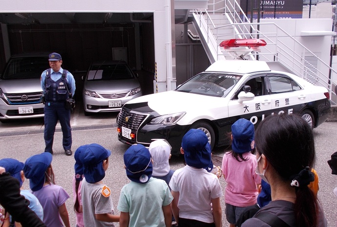 パトカーの横に立って話をしている男性警察官、整列して話を聞いている子ども達と傍で見守っている先生の写真