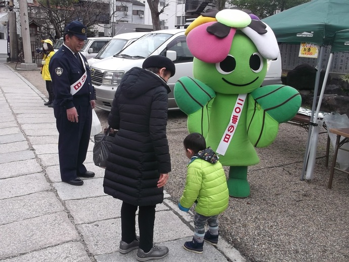ベレー帽を被った高齢者と緑色の上着を着た幼児が110番の日というたすきを掛けた東成区のキャラクターふれんじーにおじぎをしている様子を警察官の男性が見守っている写真