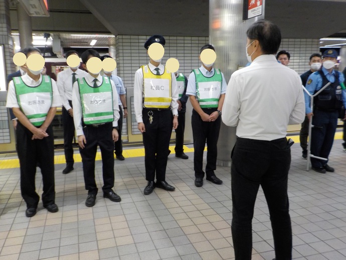 役名が書かれたビブスを身に着けた駅員や警察官が立っており、正面に立つワイシャツ姿の男性の方を向いている写真