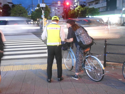 横断歩道の前で黄色の反射ベストを着用した関係者が自転車に乗っている女性にキャンペーンの説明をしている写真