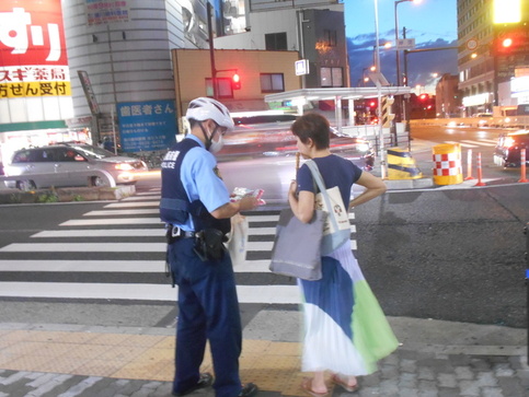 横断歩道の前で白いヘルメットを着用した制服警察官が女性にキャンペーンチラシを見せながら説明している写真