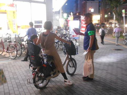 チャイルドシートが乗った自転車に乗っている親子連れに「防犯」と書かれた腕章を着用した関係者がキャンペーンチラシを持ち説明をしている写真