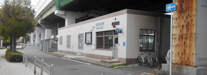 街路樹がある遊歩道近くの高架下に位置し、白壁の上下に赤茶色のアクセントカラーがあり、玄関入口に2段ほどの段差がある鶴見橋交番の外観写真