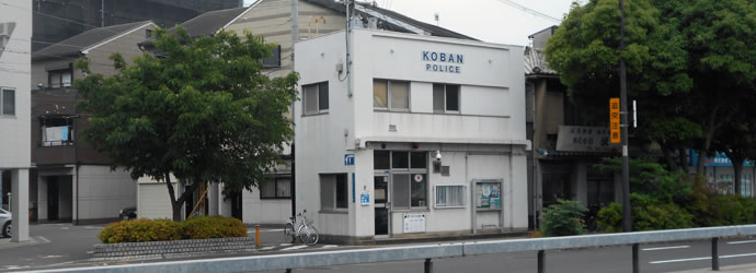 国道26号線の道路沿いにあり街路樹に囲まれた外壁が白色で2階建ての岸里南交番の外観写真