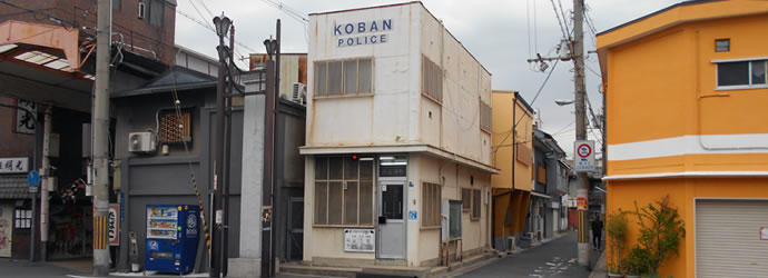 高架下すぐ近くに建てられた壁がクリーム色の陸屋根2階建ての飛田交番正面玄関前外観写真