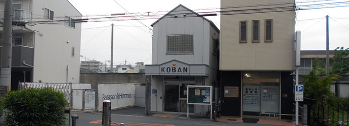 白い壁の三角屋根の2階建てで、正面右側に掲示板左側に立て看板がある天下茶屋北交番正面玄関前の外観写真