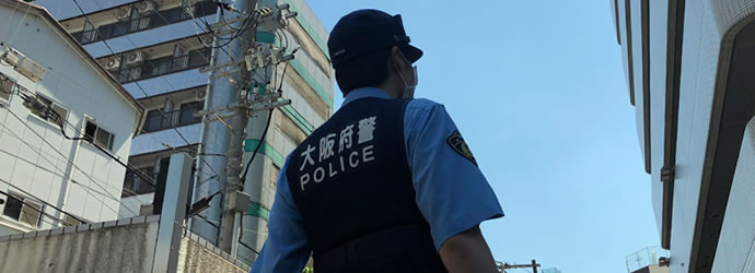 屋外で大阪府警と書かれたベストを着用した制服警察官の背中越しの写真