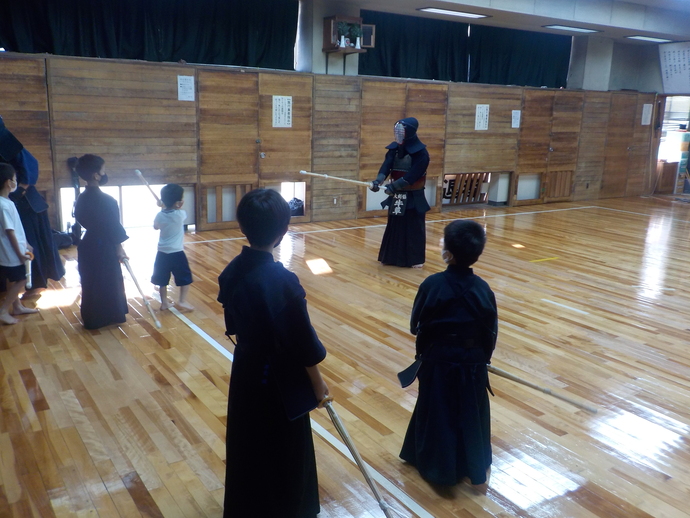 体育館内で胴着姿の剣道選手に体操服を着た男の子が竹刀で向かっていく様子を周りの子供たちが見ている写真