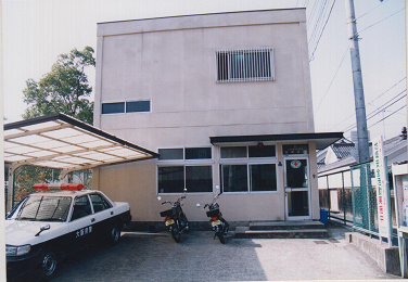 2階建てで四角形の形をしたベージュの外壁の本町交番の左前にパトカーが1台停車している写真