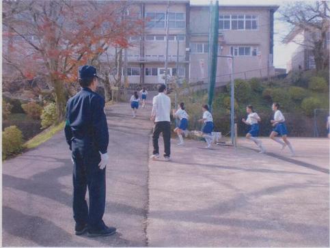 校舎前の坂道を走っている子ども達の沿道警備をしている駐在所勤務員を後方から写した写真