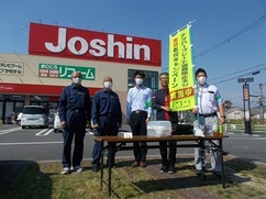 店舗前の駐車場で工具類が置かれた長テーブルの背後に全国地域安全運動の黄色いのぼり旗を持った作業服の男性とその右隣に3名の作業着姿の男性とスーツ姿の男性1名が立ち記念撮影をしている写真